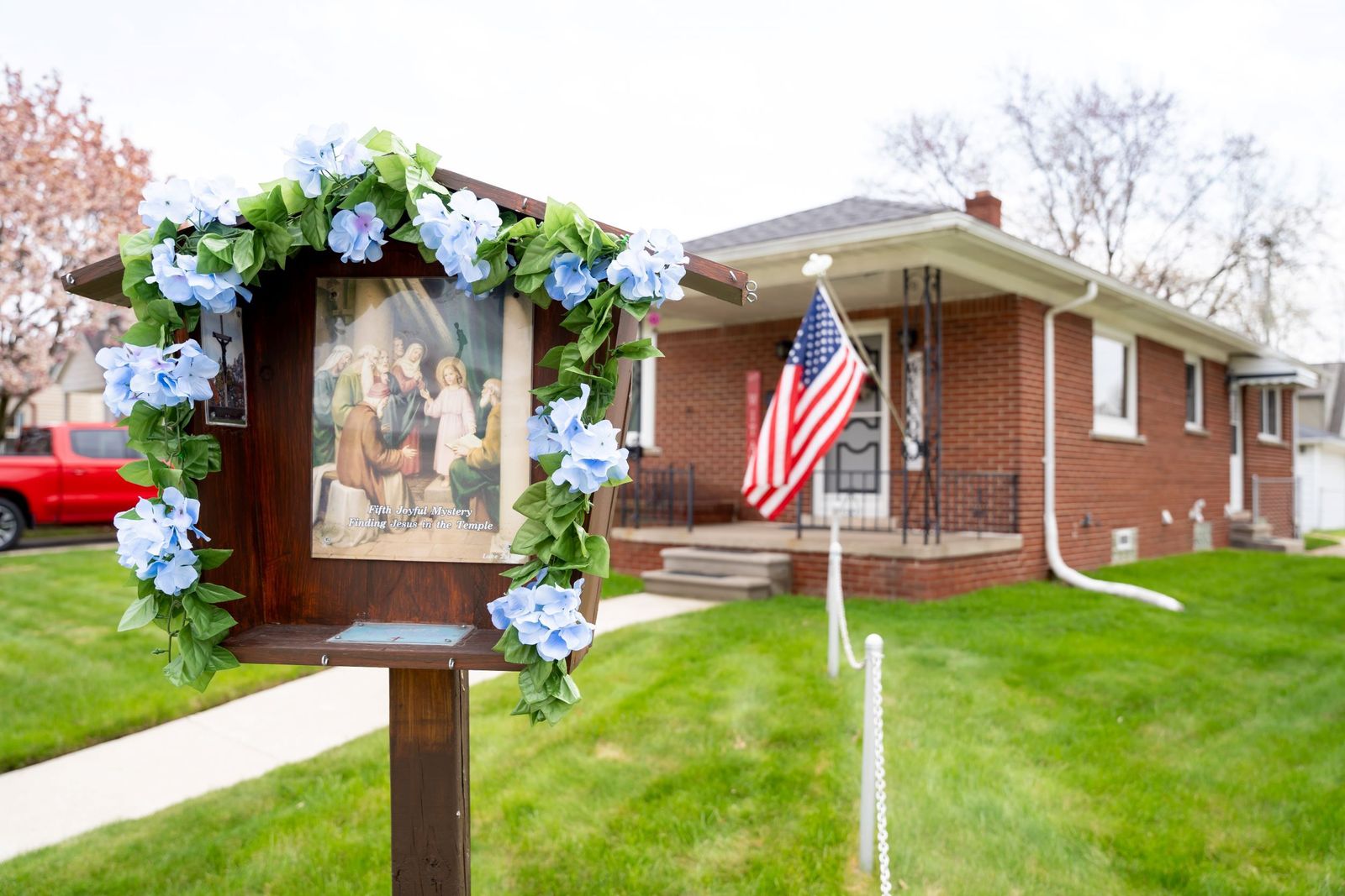 A "rosary box" dedicated to the fifth Joyful Mystery, the finding of Jesus in the temple, is pictured in a parishioner's front yard bedecked with blue flowers.